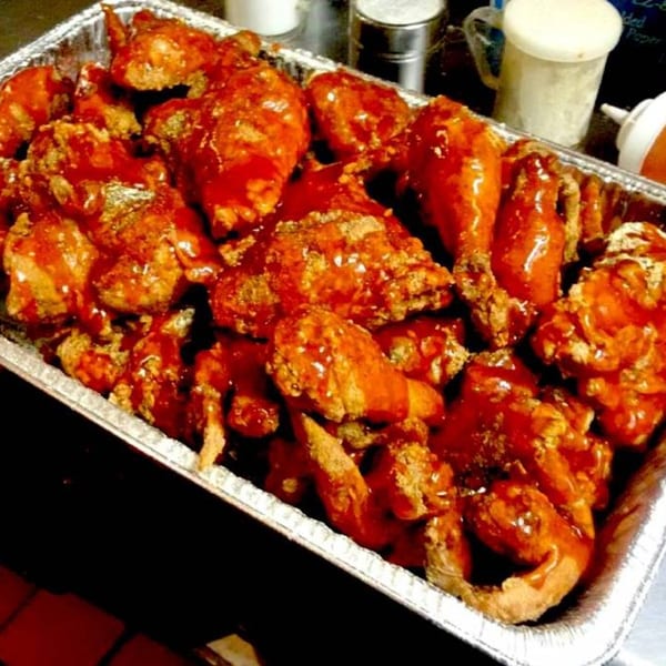 A foil pan filled with Harold's Chicken Shack's fried chicken pieces, generously coated in red sauce.